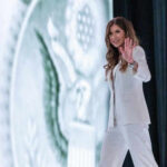 Fatal Shootings Ignite Outrage—Noem in Hot Seat Person in white suit waves on stage background