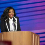 Felon Guards Congresswoman, Ends in SWAT Standoff Person in suit standing at a podium, smiling.