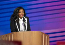 Person in suit standing at a podium, smiling.