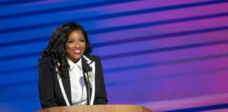 Person in suit standing at a podium, smiling.