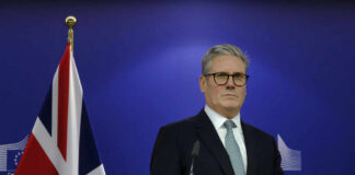 Man in suit at a podium, British flag behind.