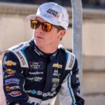Race car driver wearing sunglasses and a team uniform, sitting outdoors