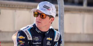Race car driver wearing sunglasses and a team uniform, sitting outdoors