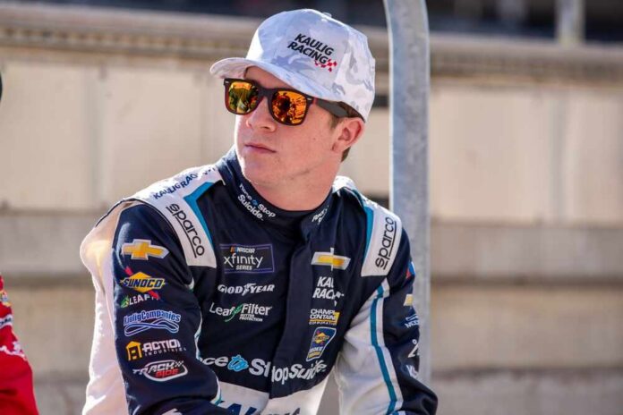Race car driver wearing sunglasses and a team uniform, sitting outdoors