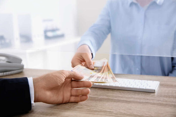 A person handing over cash through a transparent barrier at a bank counter