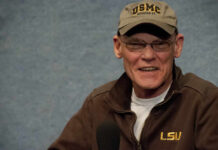 A man wearing a cap and jacket, smiling while seated in front of a blue background