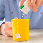 Person adding sweetener to a yellow mug of tea