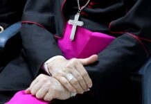 A close-up of a clergy member's hands clasped together, wearing a cross necklace and formal attire