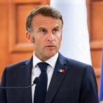 NATO Sidestepped: France Takes Lead in Hormuz A political leader speaking at a press conference with flags in the background