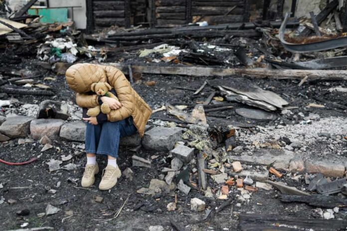 A,Girl,Next,To,A,House,Destroyed,By,The,War. A child sitting on a stone amidst a devastated landscape, holding a teddy bear