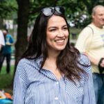 A smiling woman in a striped shirt at an outdoor event with photographers in the background
