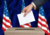 Hand placing envelope into ballot box with flag background
