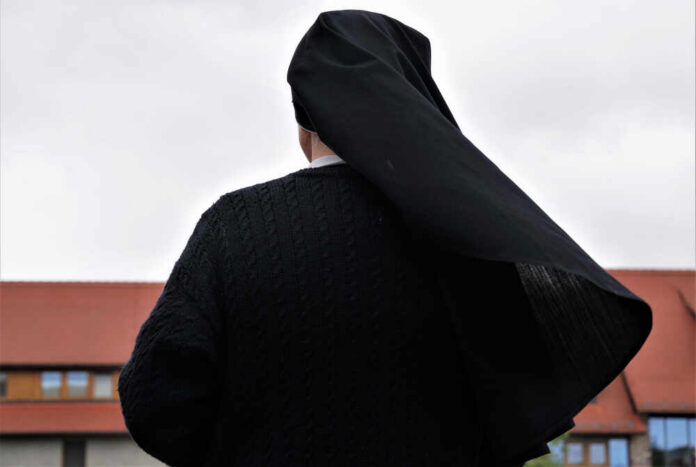 A,Christian,Nun,,Seen,From,Behind,,Looking,At,The,Old A nun in black attire standing outdoors with a cloudy sky in the background