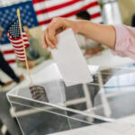 The Fight Over Voter Records Intensifies Hand placing ballot in box with American flag nearby