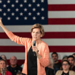 A woman speaking into a microphone with an American flag backdrop