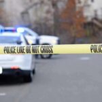 Police tape cordons off a street with patrol cars and flashing lights in the background