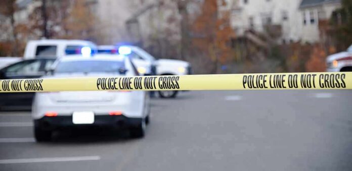 Police tape cordons off a street with patrol cars and flashing lights in the background