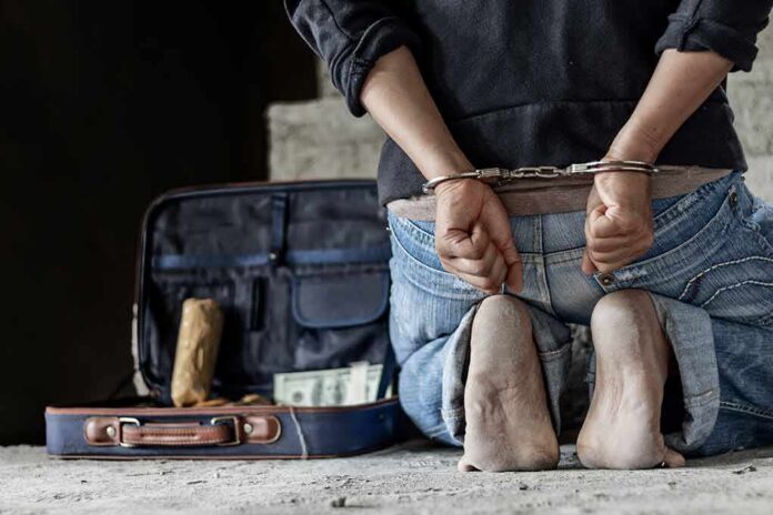 Person in handcuffs kneeling beside an open suitcase with cash