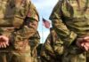 Identity Politics Gets Its Marching Orders Soldiers in camouflage stand with hands behind backs facing a U.S. flag