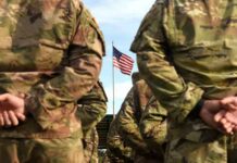 Identity Politics Gets Its Marching Orders Soldiers in camouflage stand with hands behind backs facing a U.S. flag