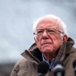 elderly man in glasses speaking outdoors into microphones