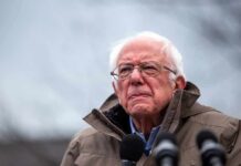 elderly man in glasses speaking outdoors into microphones