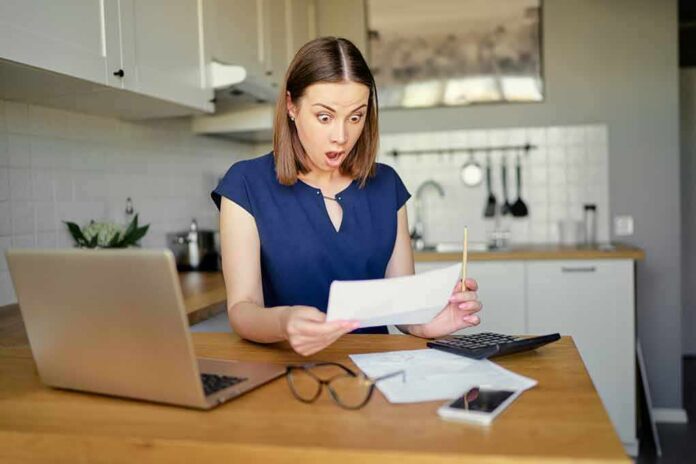 Woman in kitchen reacting to a bill at a table with laptop and calculator