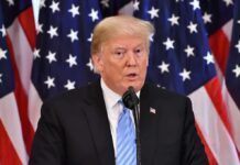 man speaking at podium with American flags behind