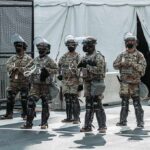 Break-Time Blowup: Guard Draws On Teammate Line of soldiers in riot gear standing on a city street