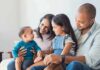 Work-First Flip: Stay-Home Parents Score Family sitting on couch with parents, daughter, and baby smiling together
