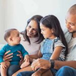 Work-First Flip: Stay-Home Parents Score Family sitting on couch with parents, daughter, and baby smiling together