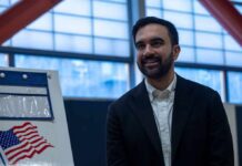 Zohran Mamdani smiling indoors near a voting sign with an American flag